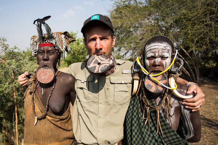 Ethiopia Tour image shows Apex Expeditions Jonathan posing with Mursi women wearing lip plates